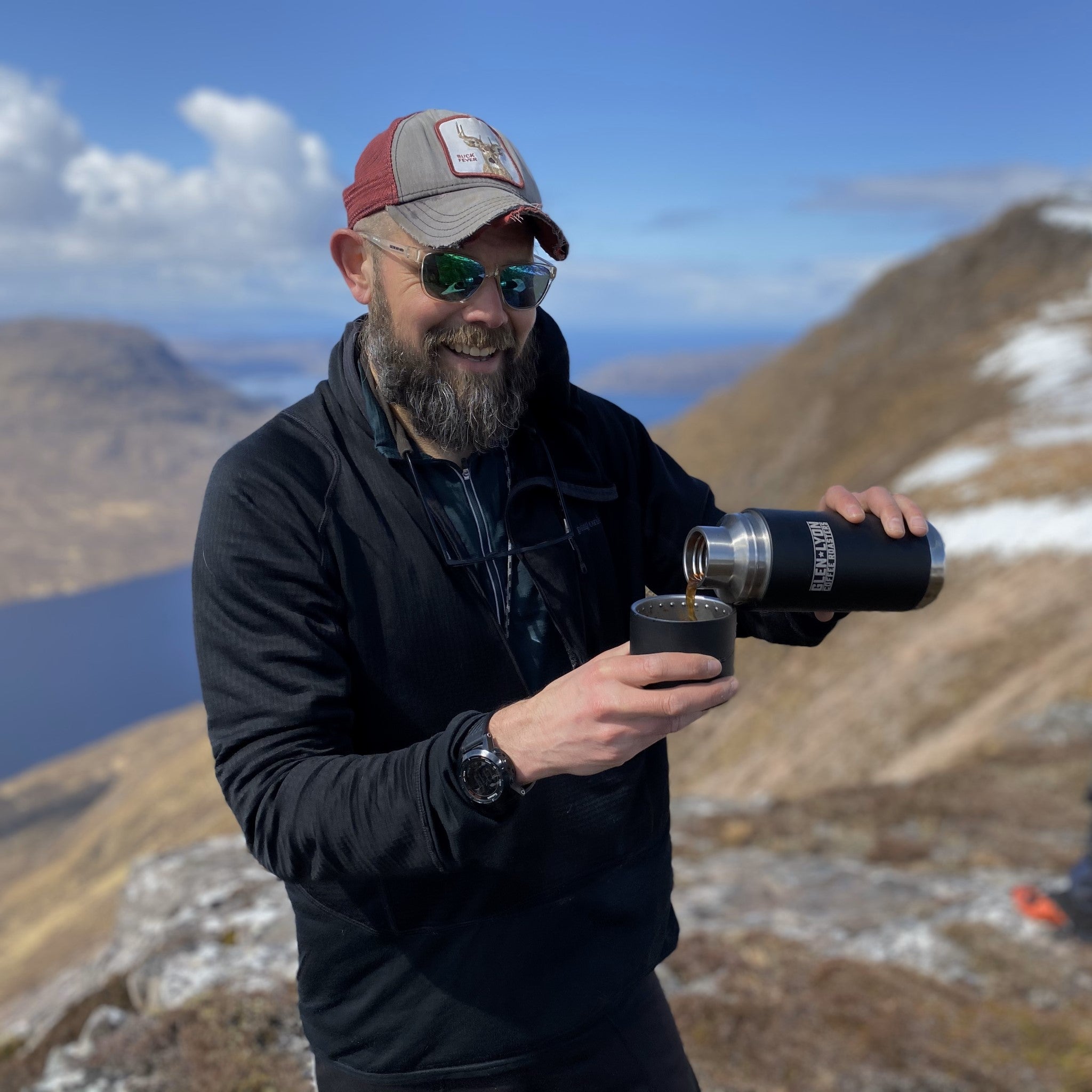 A person with a beard wearing a baseball cap on top of a hill, pouring coffee from a Glen Lyon Coffee Roasters-branded thermos into a cup