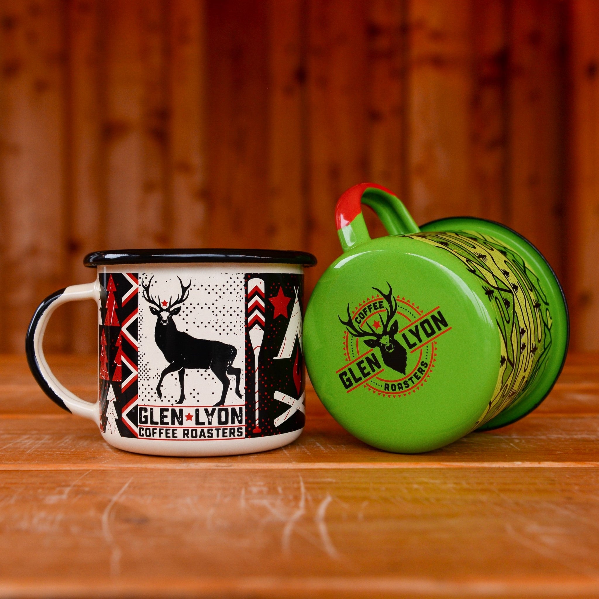 Two different coloured Glen Lyon Coffee Roasters enamel camp mugs on a wooden table, the green one on its side to reveal the Glen Lyon Coffee logo on the base