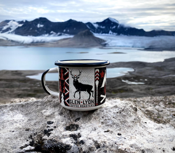 A Glen Lyon Coffee Roasters enamel mug featuring the stag logo, sitting on a glacier in the arctic with lakes and mountains behind
