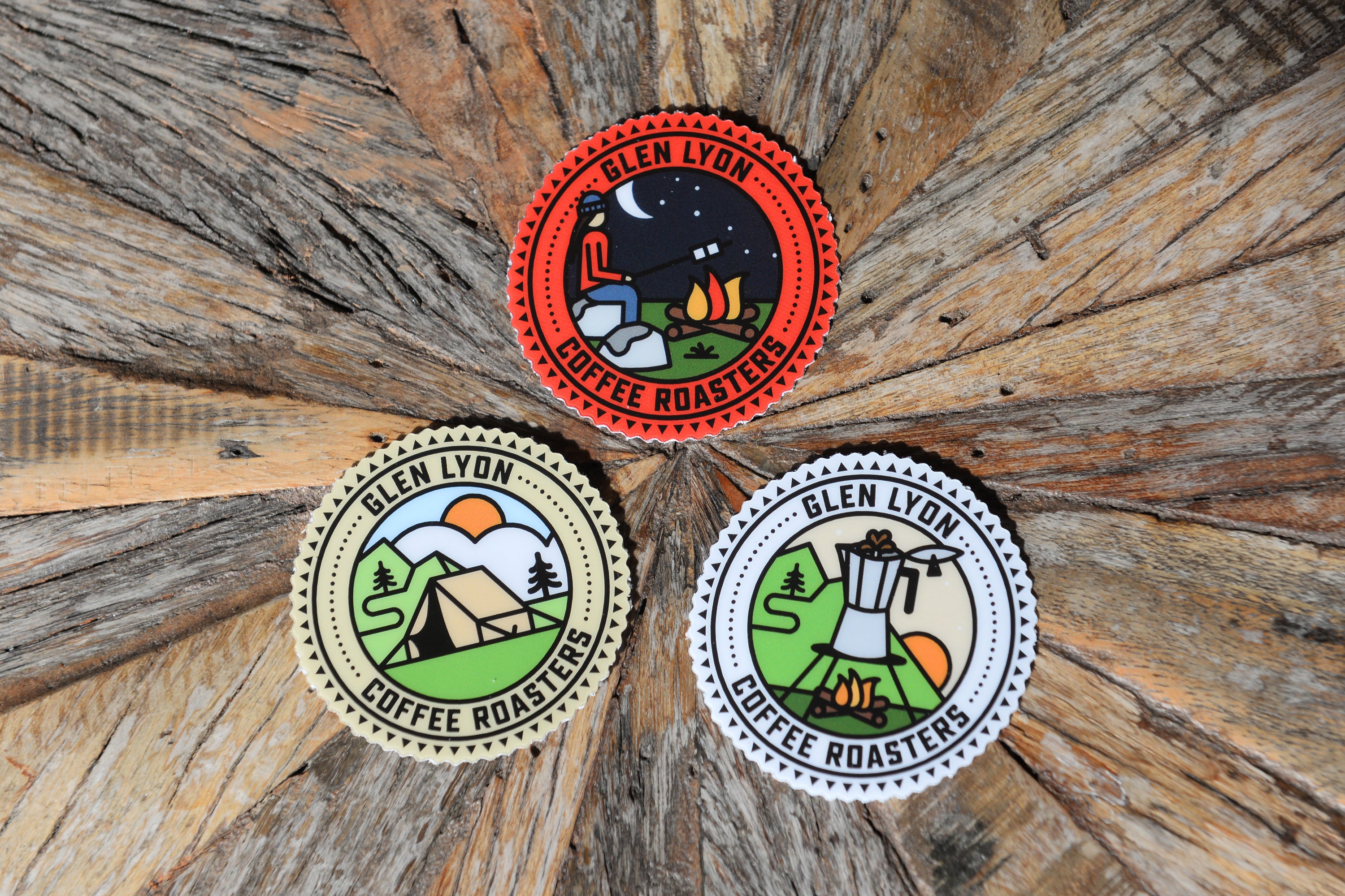 Three round Glen Lyon branded stickers with graphic scenes, placed in a triangular composition on top of a wooden background.