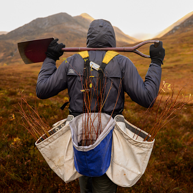 A person in waterproofs with their back to the camera walks in the Scottish Highlands holding a shovel and carrying bags full of saplings as part of Glen Lyon Coffee Roasters' annual tree planting expedition