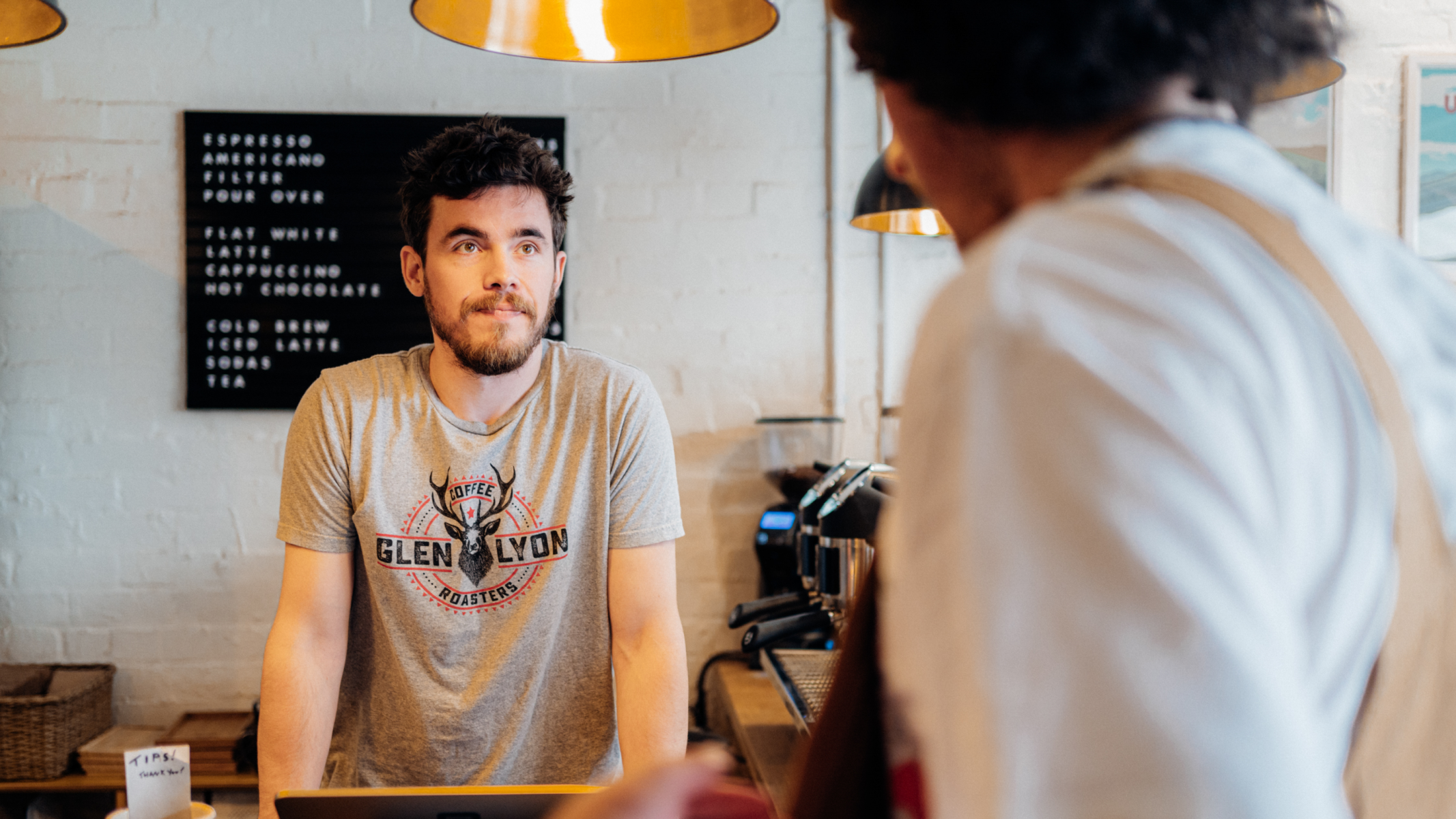 Medium shot of a customer (foreground, our of focus) speaks to a barista leaning on the bar at Glen Lyon Coffee Roasters' roastery cafe