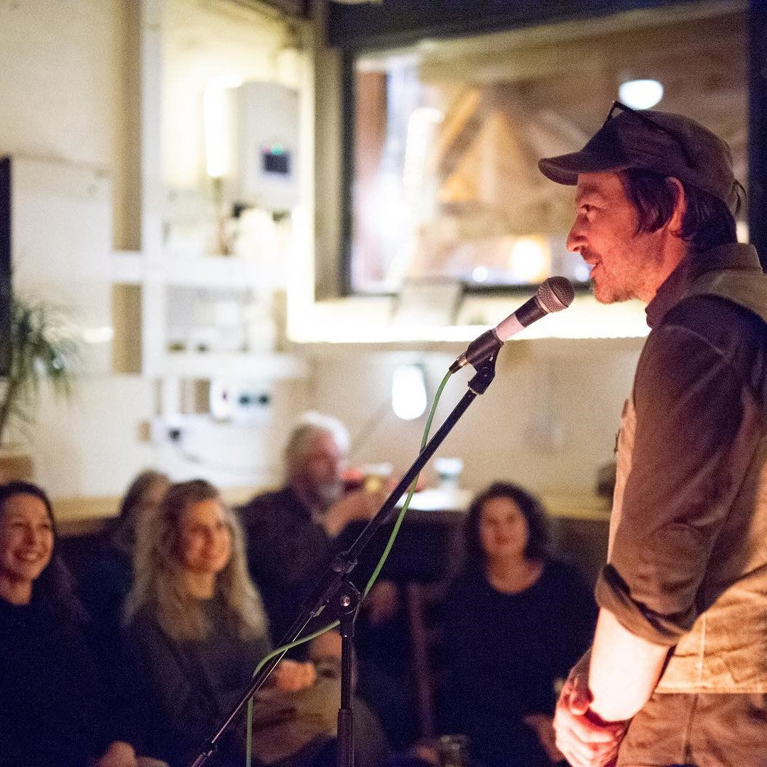 A person stands behind a microphone while seated audience members look on at a Glen Lyon Coffee Roasters Live at the Roastery poetry event