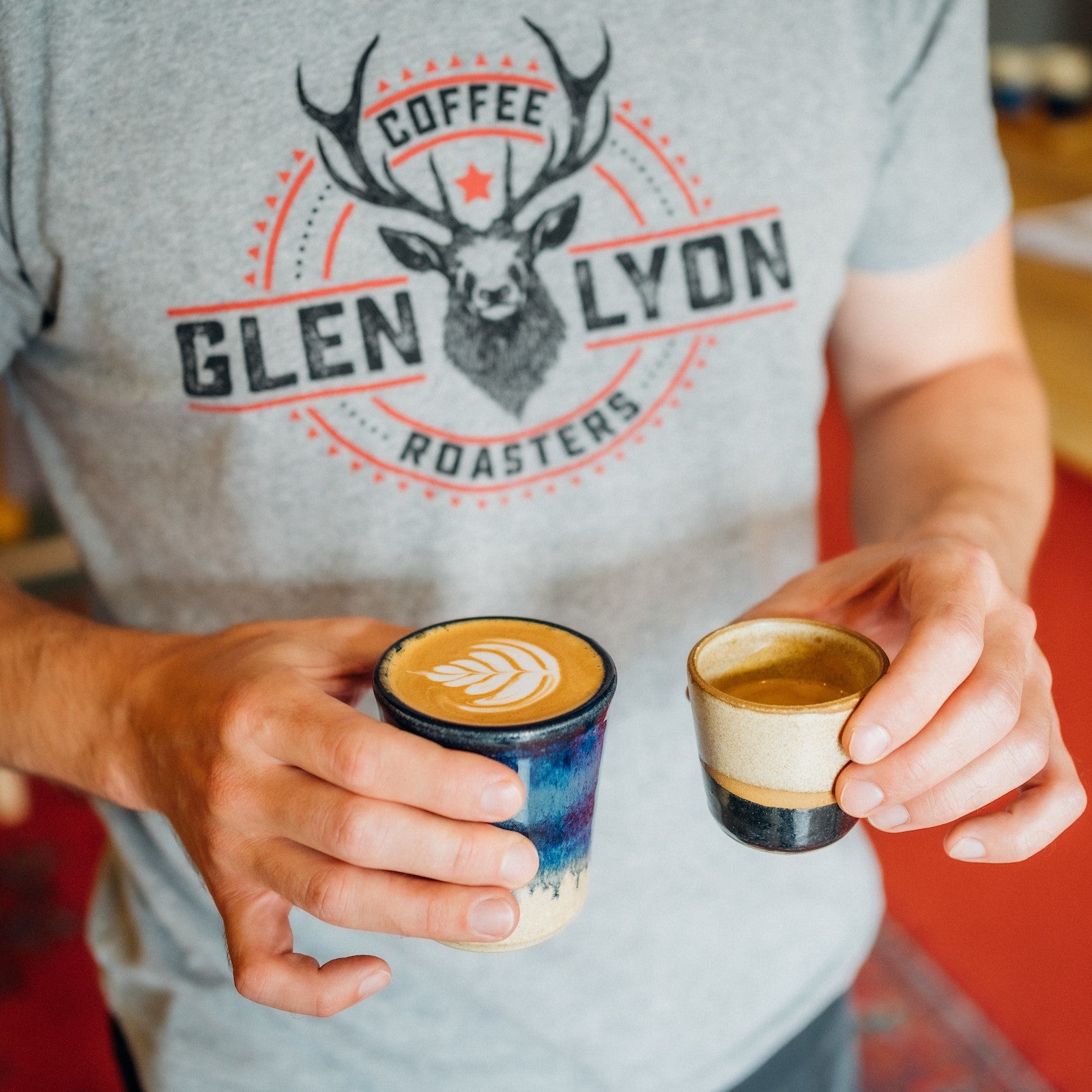 A person wearing a Glen Lyon Coffee Roasters T-Shirt carries a flat white and an espresso inside Glen Lyon Coffee Roasters roastery cafe in Aberfeldy
