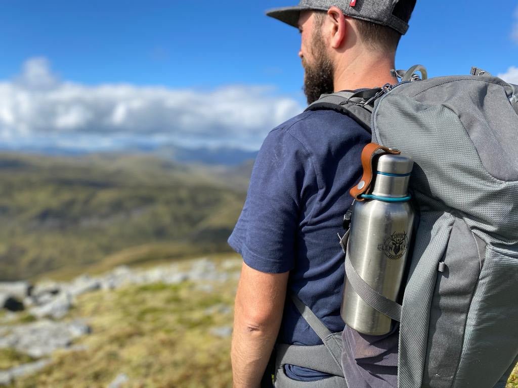 A man stands on a Scottish mountaintop wearing a rucksack with a Glen Lyon Coffee Roasters water bottle in the side.