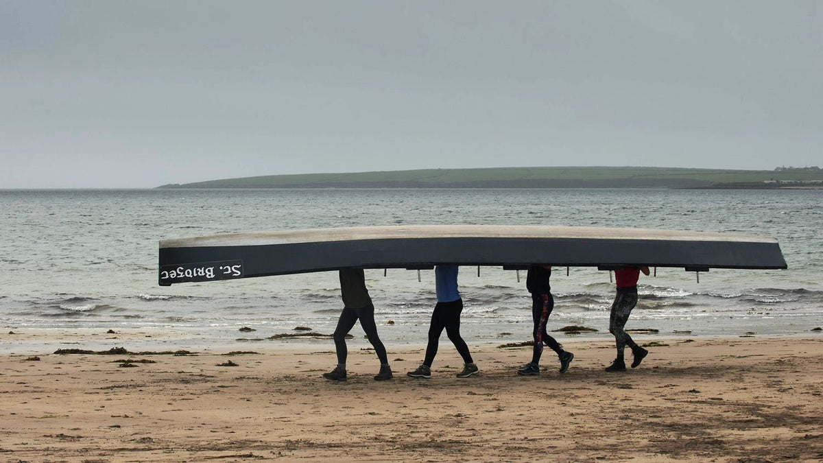 A still from the movie Tarrac, showing four people walking on a beach carrying a boat on their heads 