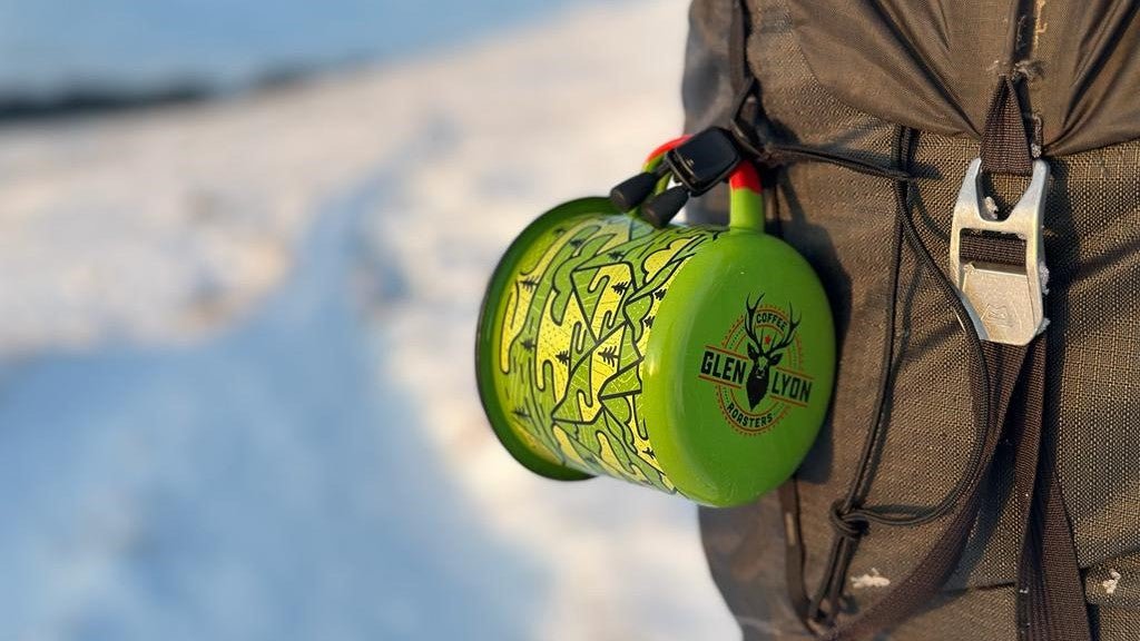 A green, yellow, and red camping mug with black mountain and tree designs hangs from the back of a camping rucksack.