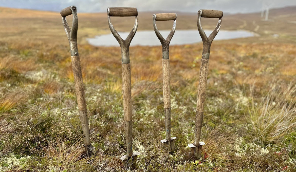 Four wooden spades are pushed into the soil in front of a beautiful Scottish lochan, resting while the tree planters do also.