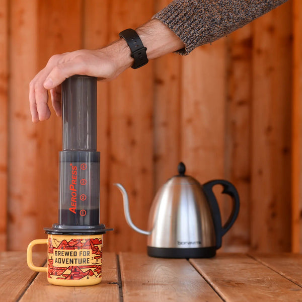 A hand pressing down on an Aeropress plunger sitting atop a 'brewed for adventure' enamel mug, with a goose neck kettle in the background.