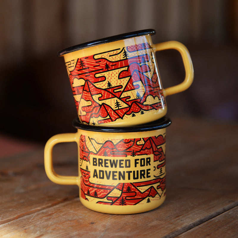 Two orange and red enameled camp mugs with black mountain design sit stacked atop one another against a wood background.