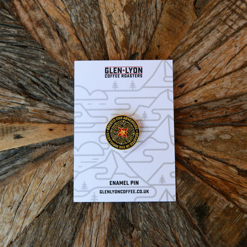 A black compass pin contrasts against a black rucksack amongst autumn foliage. It reads "Brewed for Adventure".