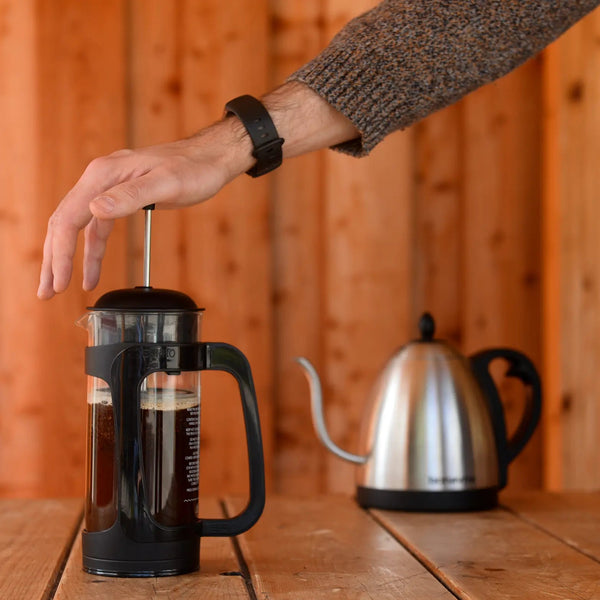 Hand pressing down on plunger of a cafetiere with brewed coffee inside, with a goose neck kettle in the background.