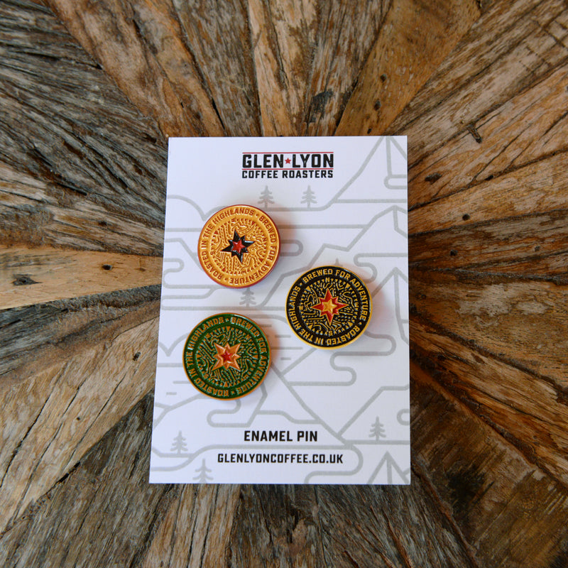 A set of three pins: a black, a yellow, and a green compass pin sit on a card on a wood table.