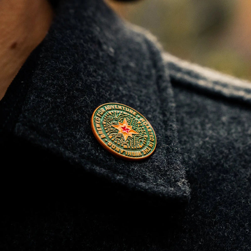 A green, gold, yellow, and red compass pin contrasts against a woolen collar of a walker.