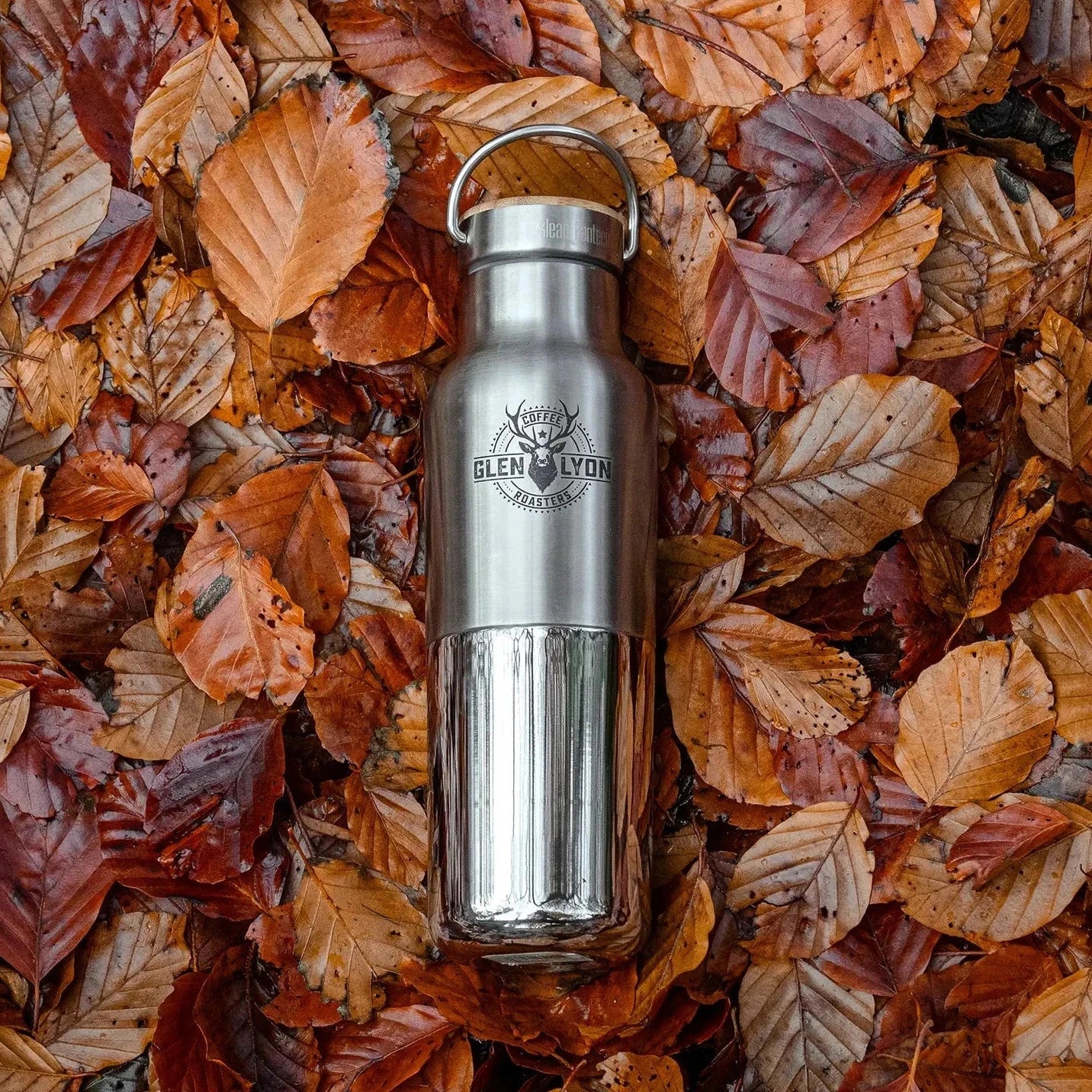 A stainless steel insulated flask lays on the forest floor. The bottom of the flask is polished and the top half is matte.