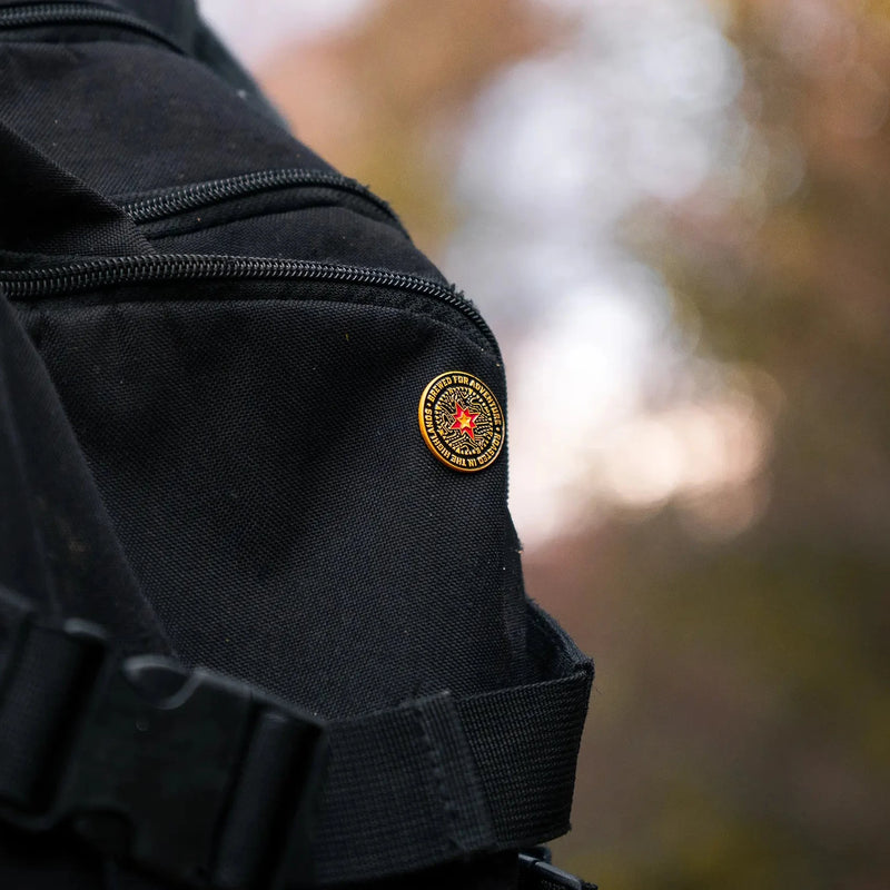 A black, gold, yellow, and red compass pin contrasts against a black rucksack amongst autumn foliage.