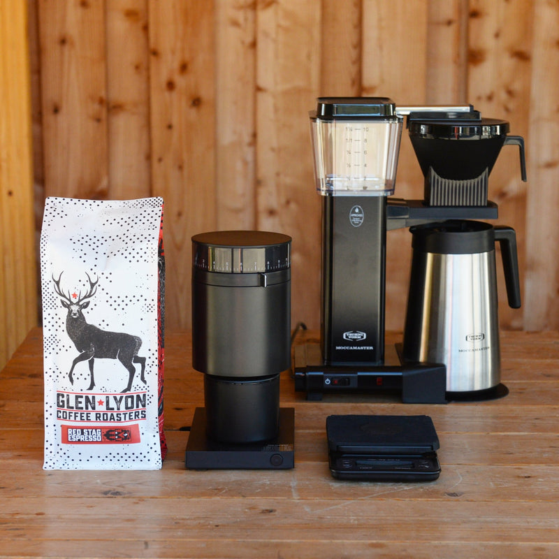 The black and stainless steel Moccamaster, black Opus, Rhino scales, and a kilo of Red Stag sit on a wooden table.