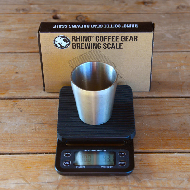 A small stainless steel cup sits atop the black Rhino brewing scales on a wooden table.
