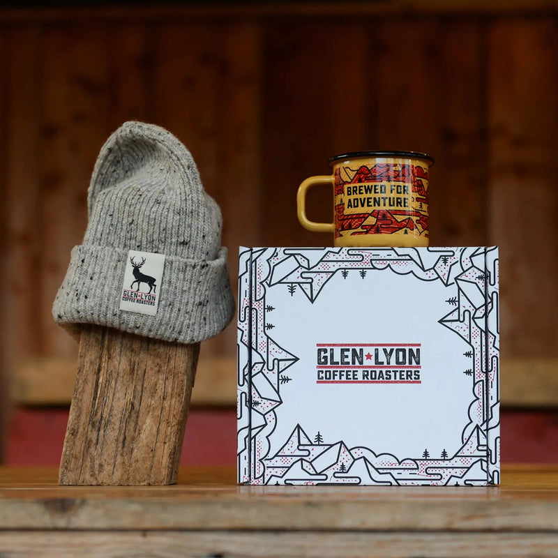 A speckled grey Scottish wool hat with Glen Lyon Coffee logo and an orange and red enameled camp mug sit atop a branded box.