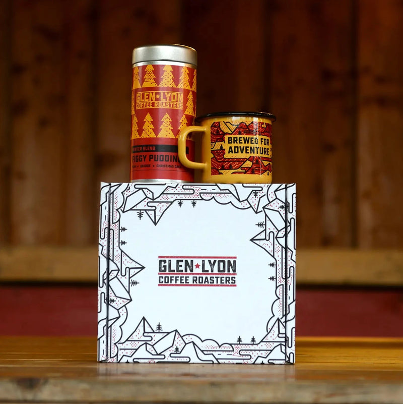 A metal 220g tin of Figgy Pudding and an orange and red enameled camp mug with black mountain design rest atop a branded box.