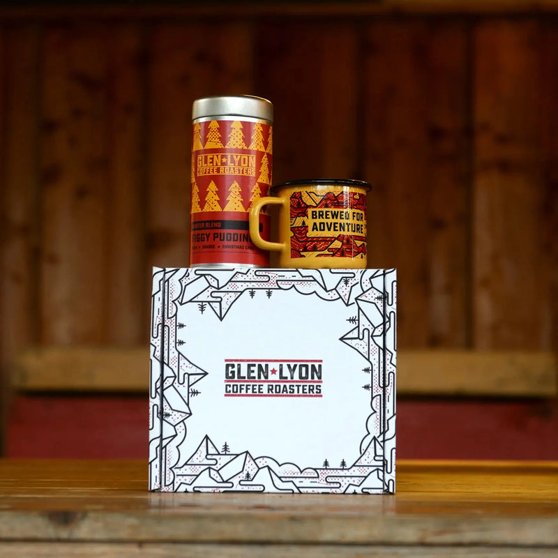 A metal 220g tin of Figgy Pudding and an orange and red enameled camp mug with black mountain design rest atop a branded box.