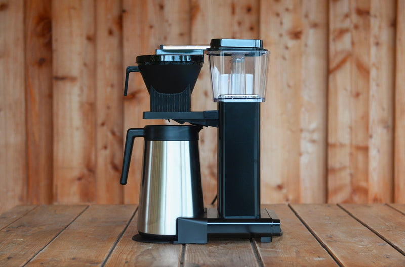 The black and stainless steel Moccamaster sits on a wooden table. The insulated carafe is polished steel.