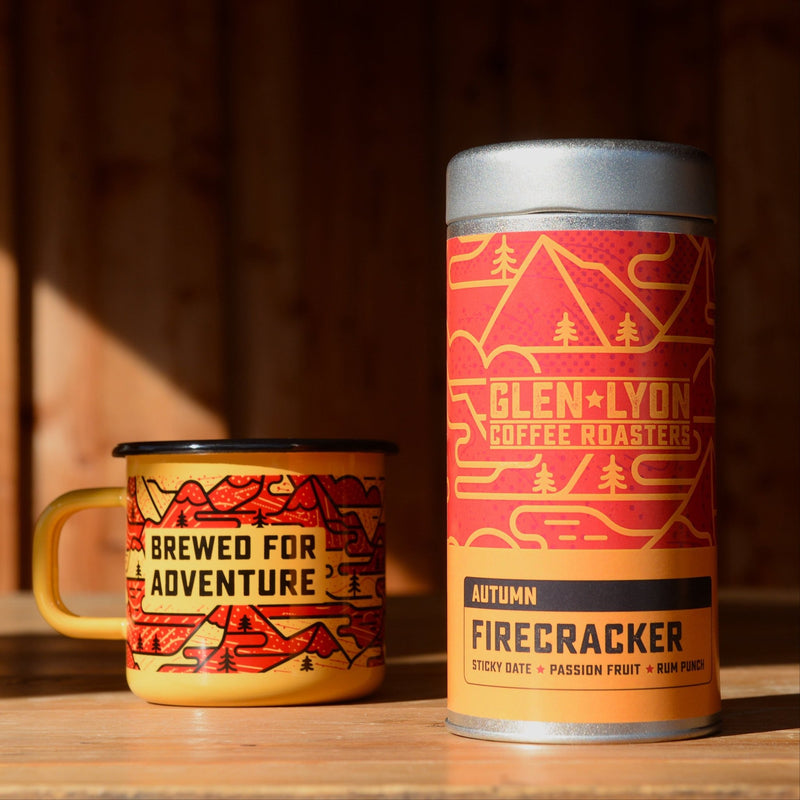 A metal 220g tin of Figgy Pudding and an orange and red enameled camp mug with black mountain design rest atop a wood table.