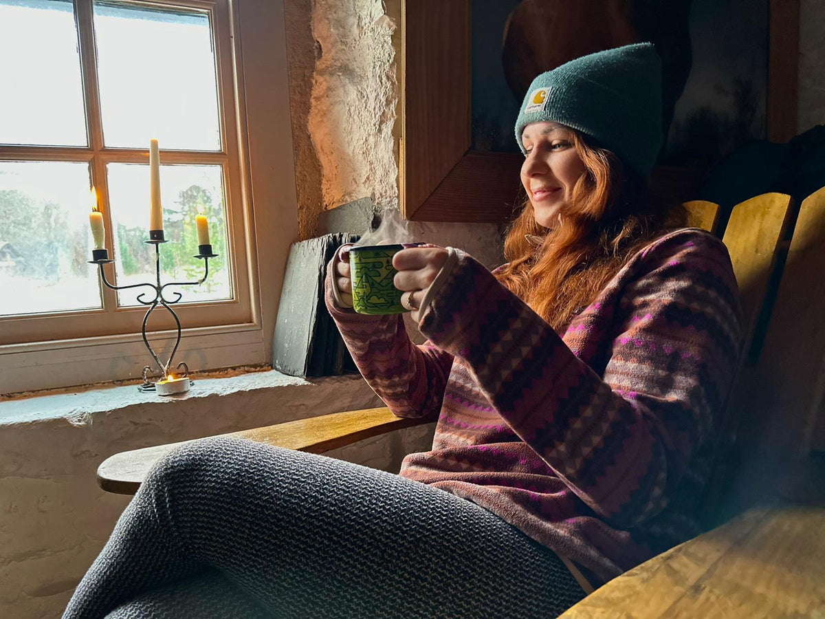 Medium shot of a woman in a beanie and jumper sitting in a bothy holding a Glen Lyon Coffee enamel camp mug