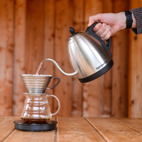 Hand pouring boiled water from a goose neck kettle into a kalita wave, sitting atop a glass jug on a scale.