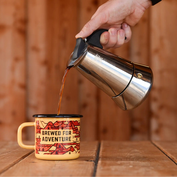 Hand pouring brewed coffee from silver moka pot into a 'brewed for adventure' enamel mug.