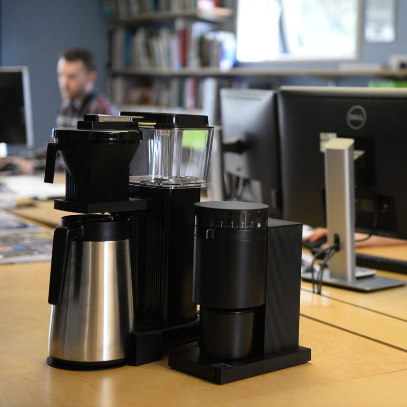 The black and stainless steel Moccamaster and black Opus sit on an office counter with an office worker in the background.