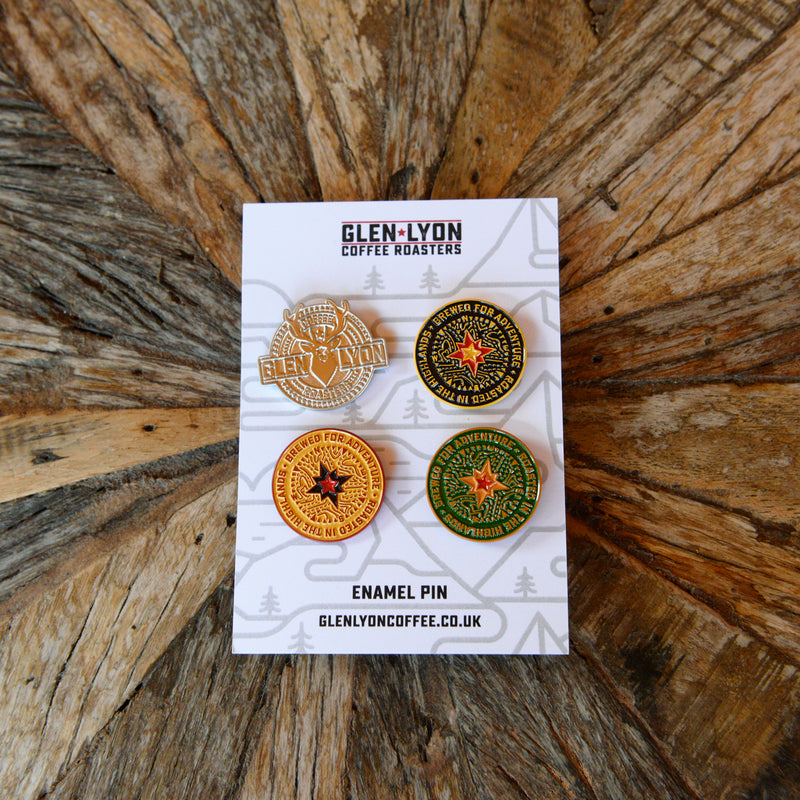 A set of four pins: silver Glen Lyon stag logo and a black, a yellow, and a green compass pin sit on a card on a wood table.