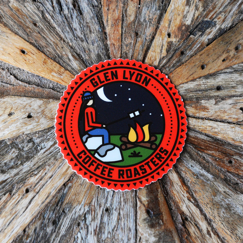 Red coloured round sticker with a graphic scene of a person roasting marshmallows on an open fire at night, with 'Glen Lyon Coffee Roasters' text printed around the scene. Sticker is placed on top of a wooden background.