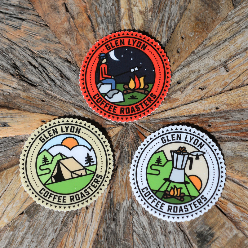 Three round Glen Lyon branded stickers with graphic scenes, placed in a triangular composition on top of a wooden background.