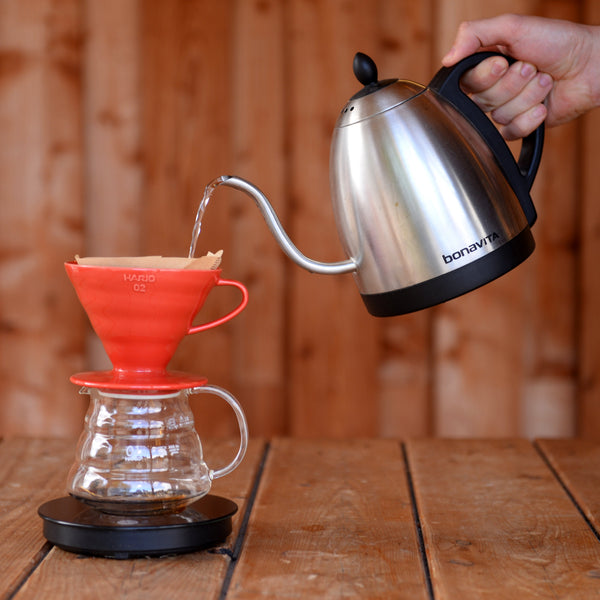 Hand pouring boiled water from goose neck kettle into a red Hario V60 atop a glass jug, sitting on a scale.