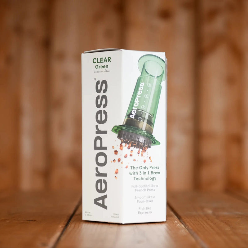 A boxed, green Aeropress sits on a wooden table. The box shows a translucent green Aeropress with droplets of coffee.