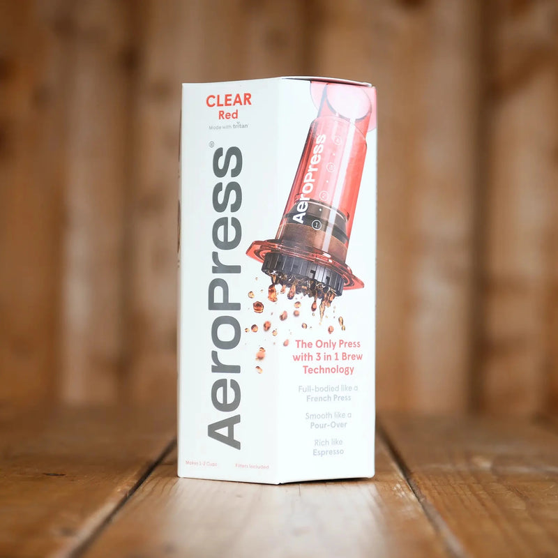 A boxed, red Aeropress sits on a wooden table. The box shows a translucent red Aeropress with droplets of coffee coming out.