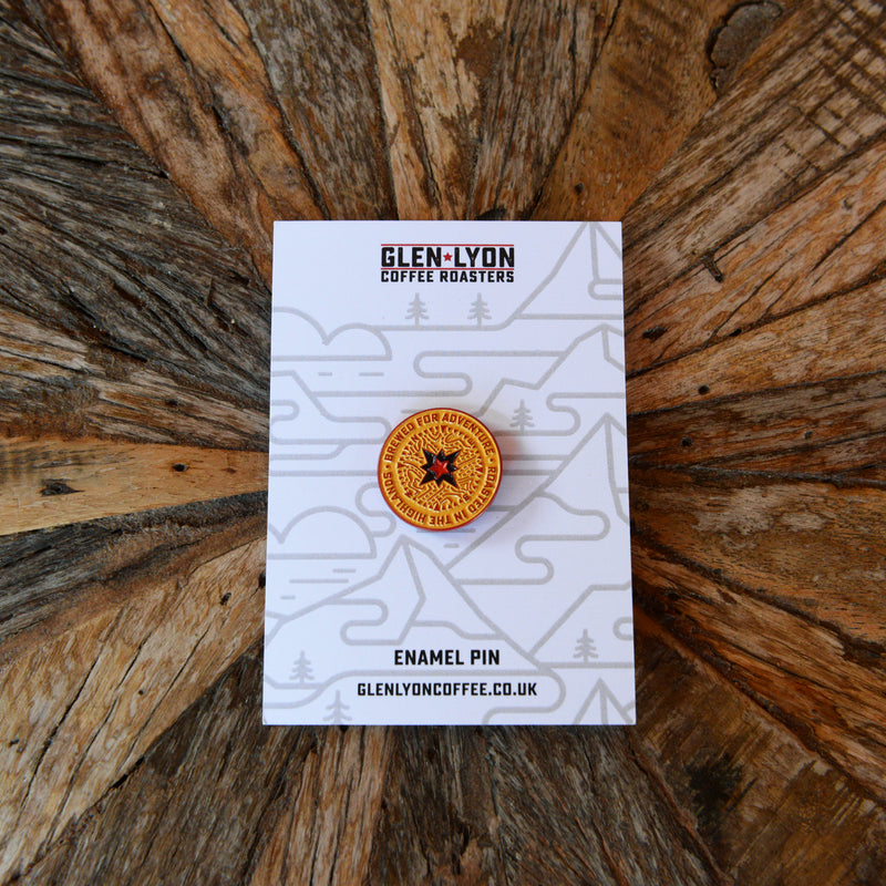 A yellow compass pin contrasts against a black rucksack amongst autumn foliage. It reads "Brewed for Adventure".