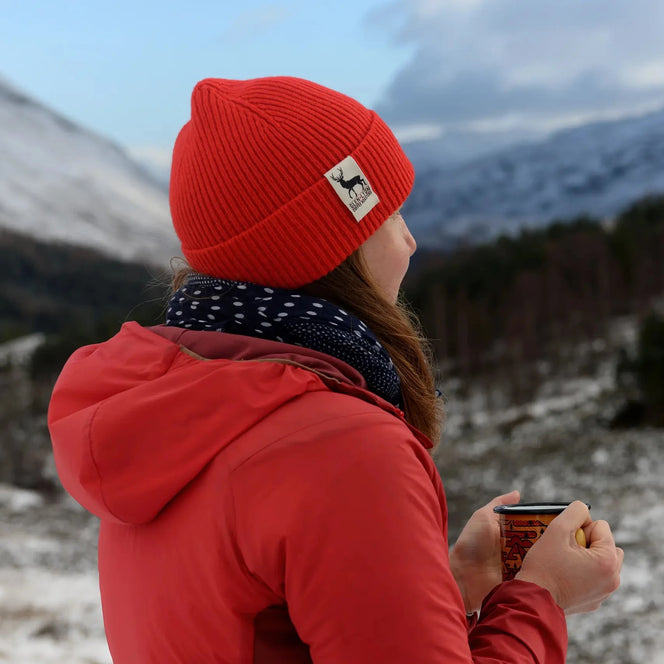 A woman in a red wool Tay Beanie stands on a Scottish mountaintop and looks out over a frozen landscape with a cup of coffee.