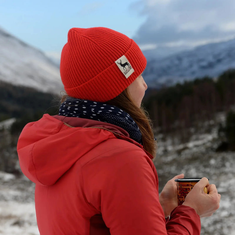 A woman in a red wool Tay Beanie stands on a Scottish mountaintop and looks out over a frozen landscape with a cup of coffee.