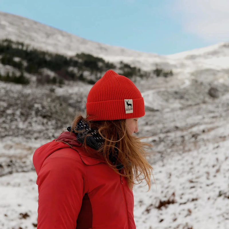 A woman in a red wool Tay Beanie stands on a Scottish mountaintop and looks down at the snow.