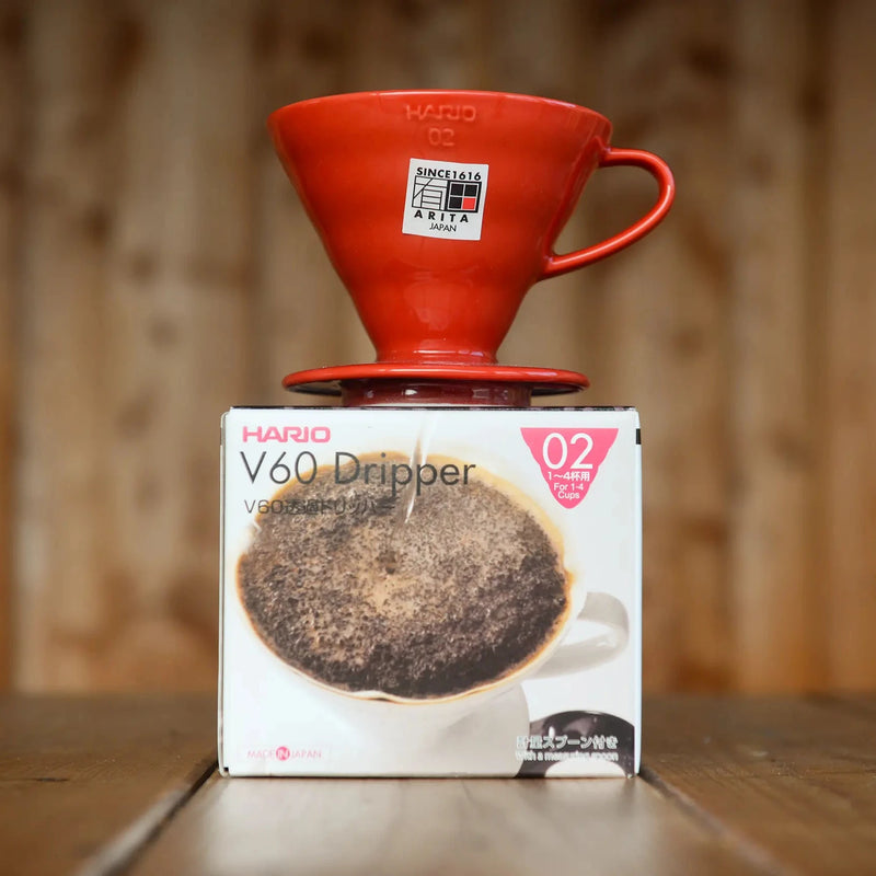 A red ceramic Hario V60 brewer with handle sits atop its cardboard box. The red glaze has a shiny, candy-like finish.