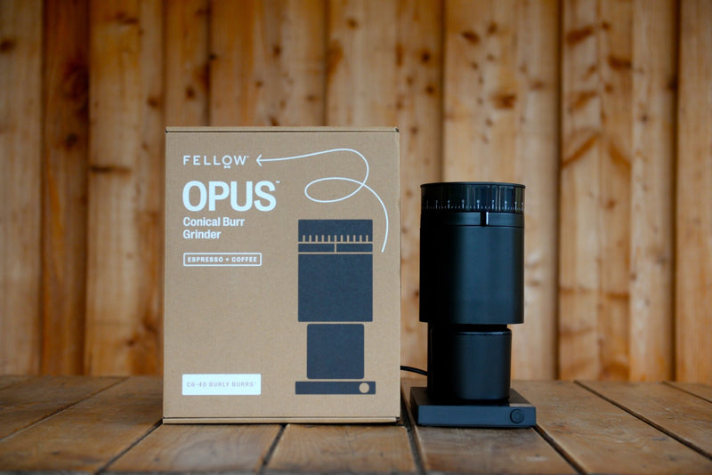 The black Fellow Opus sits on a wooden table next to its cardboard box. The design is elegant and simple with flowing lines.