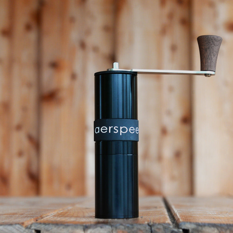 A shiny black metal Aerspeed hand coffee grinder sits on a wooden table. The handle is shiny silver steel and wood.