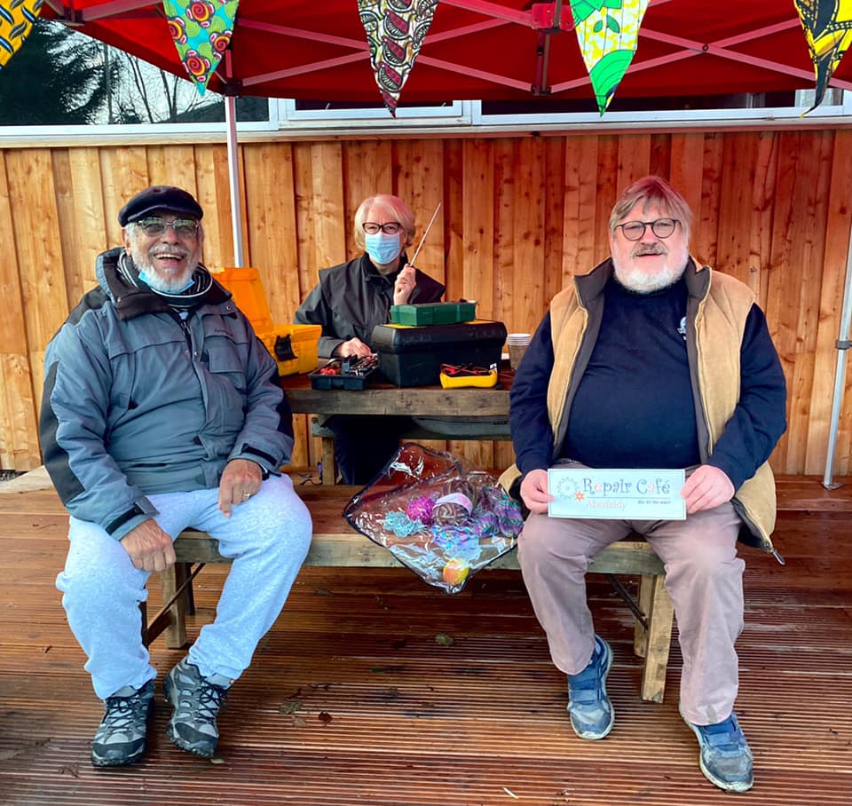 Three people smile at the camera sitting at a table under the canvas yurt outside Glen Lyon Coffee's roastery cafe during the monthly Repair Cafe